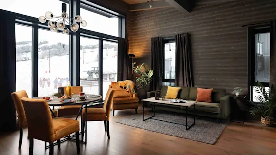 En hyggelig stue med bløde orange stole omkring et spisebord og en grøn sofa ved et vindue. Sneklædt bjerglandskab er synligt udenfor gennem store vinduer.