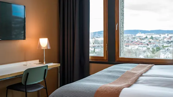 A hotel room features a bed with a gray and brown cover, a wall-mounted TV, and a desk with a lamp and chair. Large window reveals city and mountain view.