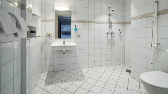 A white tiled bathroom features a wall-mounted sink, mirror, and walk-in shower with grab bars. Towels and a hairdryer are attached to the wall. A distant room is visible.