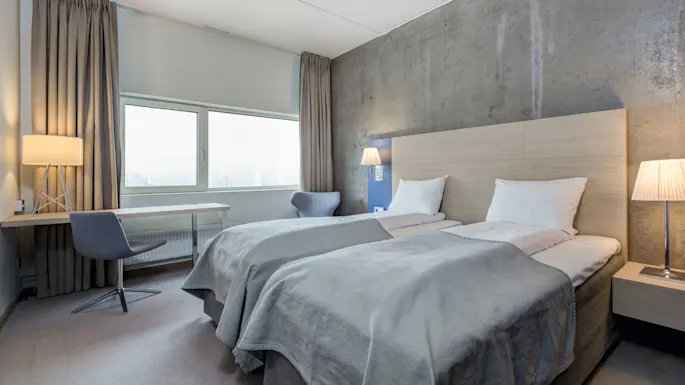 Two neatly made twin beds with white linens and gray blankets are in a minimalist room. A lamp and chair sit beside a window, with light gray curtains.