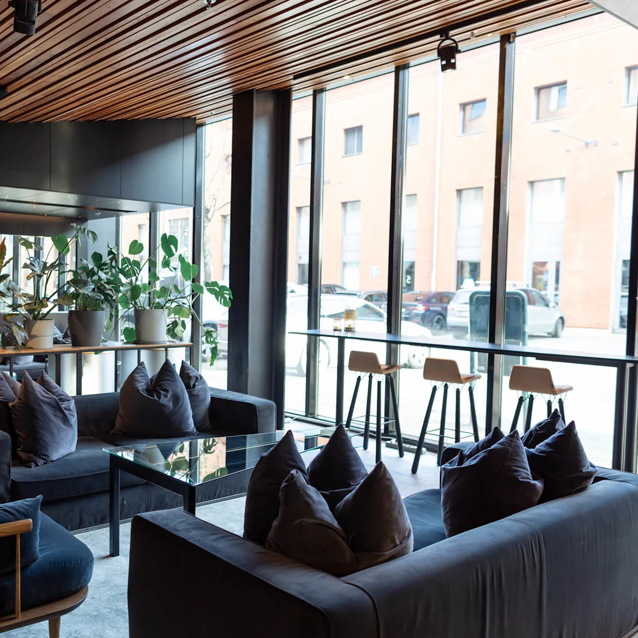 Sofas adorned with plush pillows, set in a modern lounge with wooden ceiling, surrounded by indoor plants and large windows overlooking a street scene with parked cars.