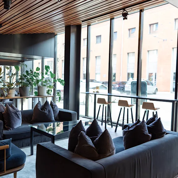 Soffor dekorerade med mjuka kuddar, placerade i en modern lounge med trätak, omgivna av inomhusväxter och stora fönster med utsikt över en gatuscen med parkerade bilar.