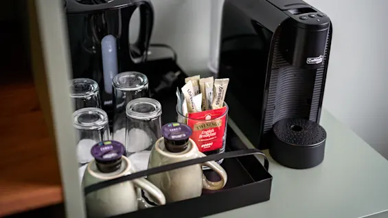Vattenkokare och kaffemaskin står på en bänk bredvid glas, muggar, tepåsar och kaffekapslar. Bänken är en del av ett hotellrumsförfriskningsuppsättning.