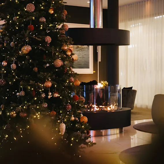 Et pyntet juletre med glitrende lys ved siden av en moderne peis i en koselig hotellobby.