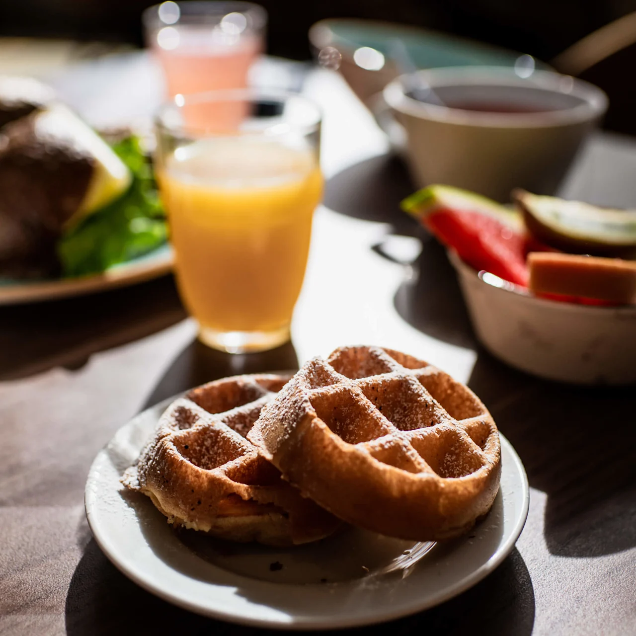 Waffles on a white plate dusted with powdered sugar. Orange juice and various breakfast items in the background, all set on a wooden table with sunlight streaming in.