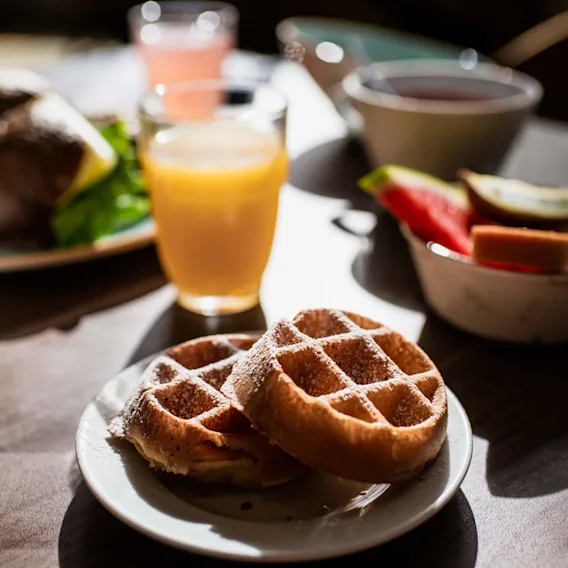 Våfflor på en vit tallrik pudrade med florsocker. Apelsinjuice och olika frukostartiklar i bakgrunden, allt placerat på ett träbord med solljus som strömmar in.