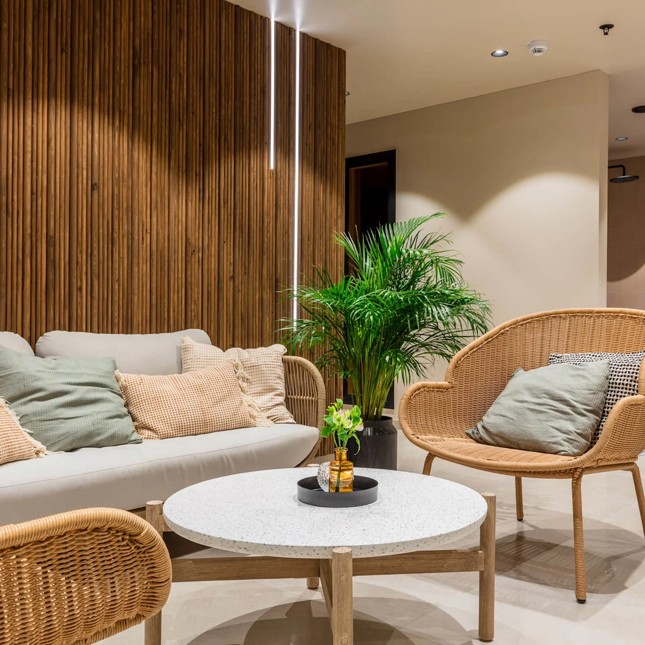 A modern lounge area with rattan chairs, a sofa with cushions, and a plant. The setting is a chic, well-lit space with wooden paneling and tiled flooring.