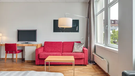 Punainen sohva sijaitsee modernissa huoneessa puulattialla, seuranaan suorakulmainen sohvapöytä. Suuri ikkuna tuo tilaan luonnonvaloa, avautuen ulkonäkymälle.