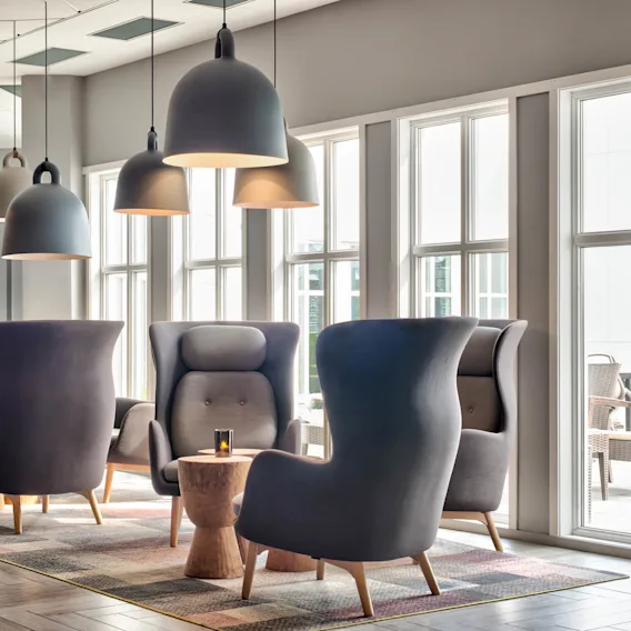 Grå lenestoler sitter rundt et trebord, opplyst av pendellamper i et lyst, moderne rom med store vinduer og tregulv.