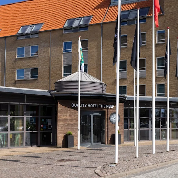 Hotellets indgang med skiltet "QUALITY HOTEL THE REEF" er omgivet af høje flagstænger. Bygningen har flere vinduer, et orange tegltag og en klar blå himmel i baggrunden.