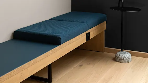En väggmonterad TV, inaktiv, vetter mot en liten, blå vadderad bänk. I närheten står ett svart sidobord med marmorbas på ett trägolv i ett modernt, minimalistiskt rum.