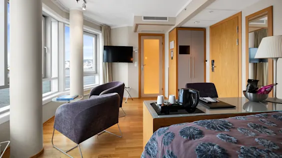 Et koselig hotellrom har to moderne stoler ved store hjørnevinduer med utsikt over havet. En veggmontert TV peker mot en seng med et blomstermønstret sengeteppe. Snacks ligger på et bord.
