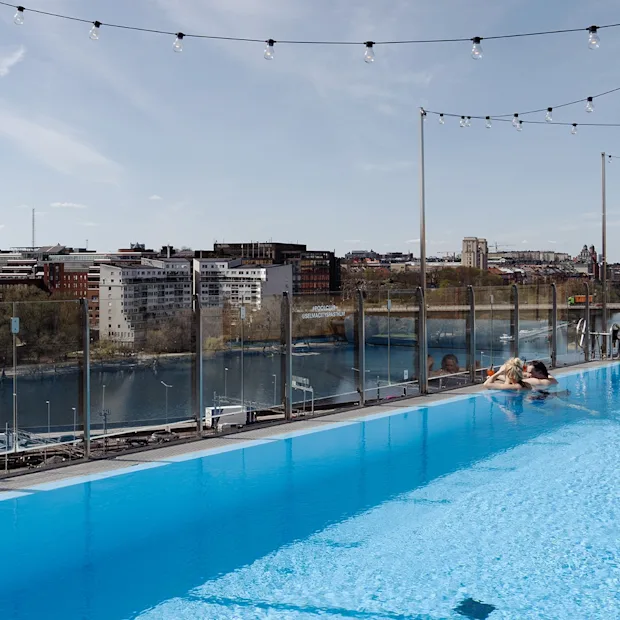 Et basseng på taket med to personer som slapper av ved kanten, omgitt av lysslynger. Utsikt over en bylandskap med bygninger og en elv under en klar himmel.