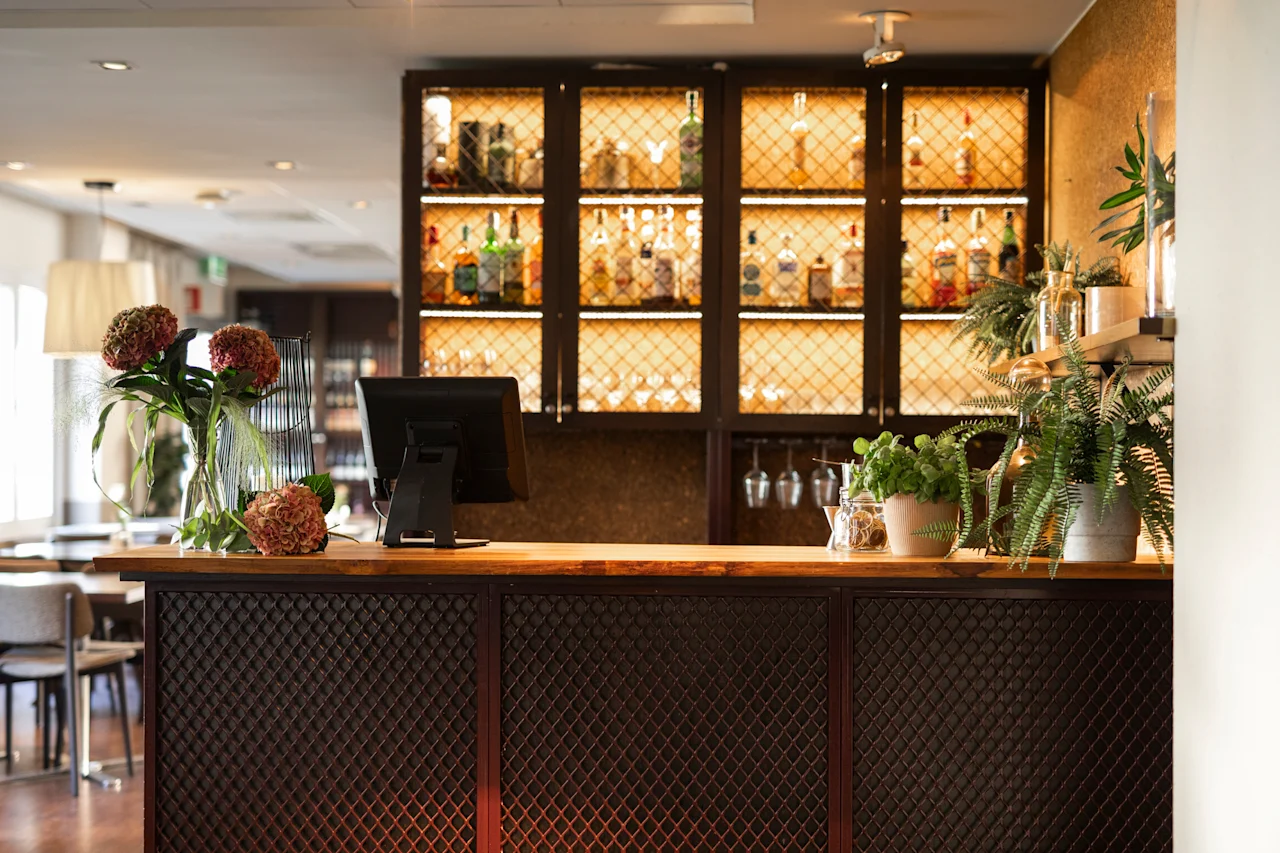 En välkomnande bar- eller receptionsdisk med en skärm, blommor och växter, mot upplysta hyllor med olika flaskor.