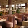 Komfortable, buede båser med glass og blomster står i en varmt opplyst restaurant med store vinduer, innendørs planter og hengende lys, og skaper en koselig spiseatmosfære.