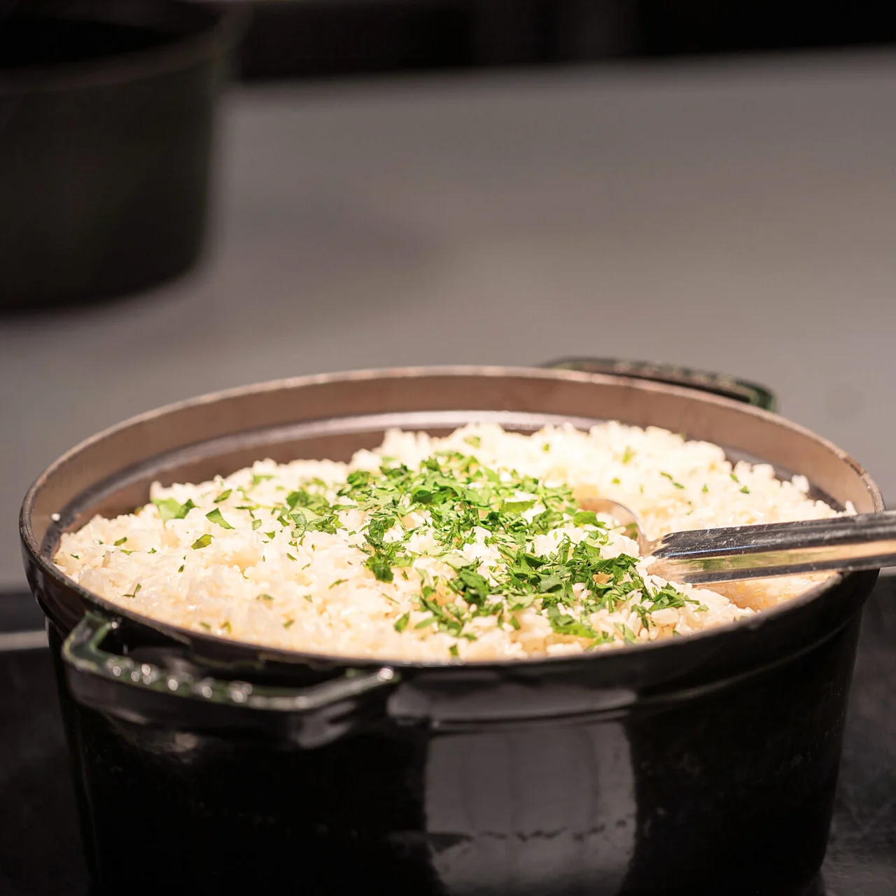 A pot holds cooked rice garnished with chopped herbs, accompanied by a metal serving spoon. It rests on a dark countertop, with soft lighting highlighting the dish.