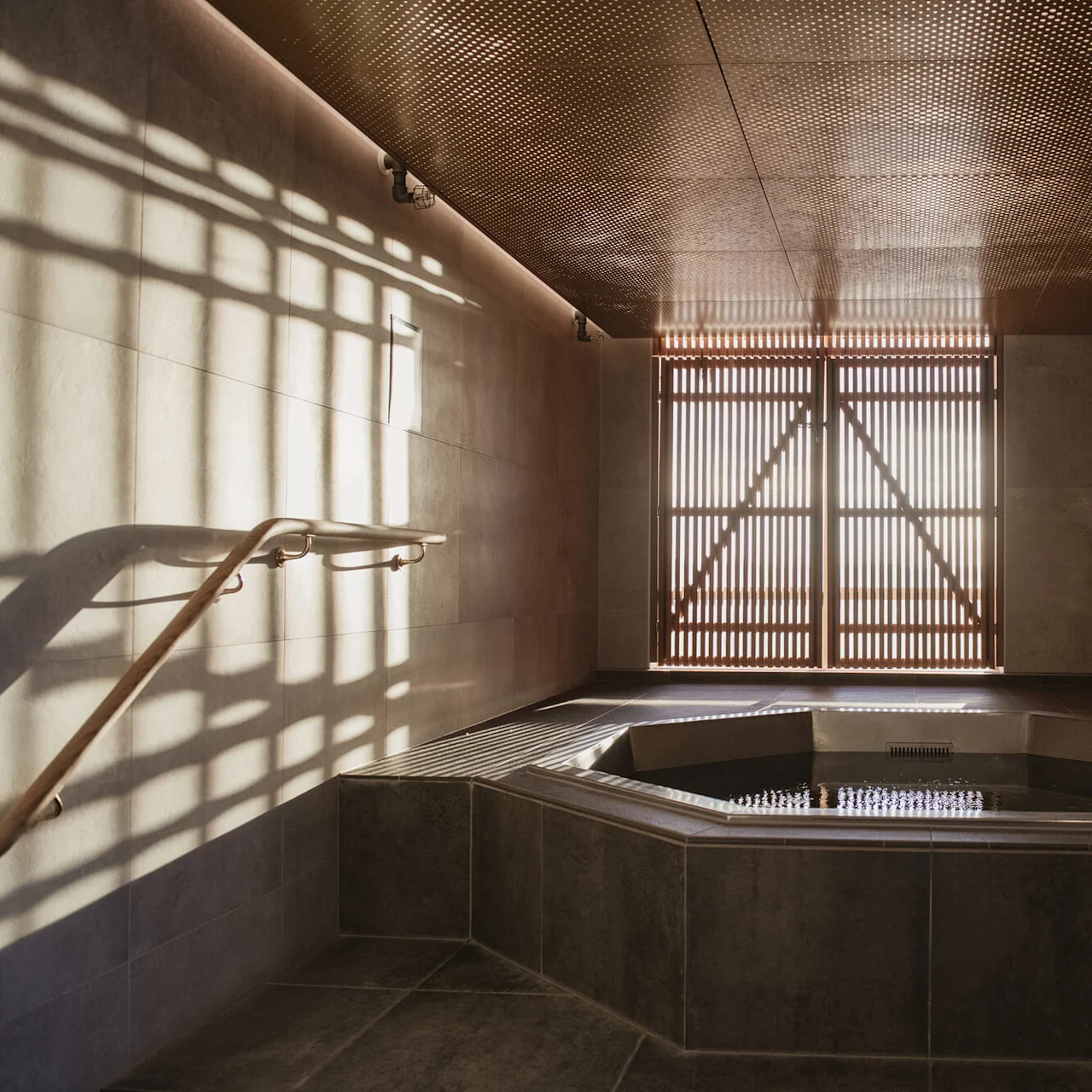 A sunlit spa room with a hexagonal hot tub is recessed into the floor, surrounded by stone tiles. A wooden screen diffuses light, casting shadows on the beige walls.