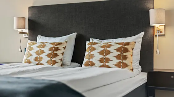 En mysig säng på Quality Hotel Ulstein med en mörk sänggavel, vita lakan och mönstrade kuddar, upplyst av sänglampor.