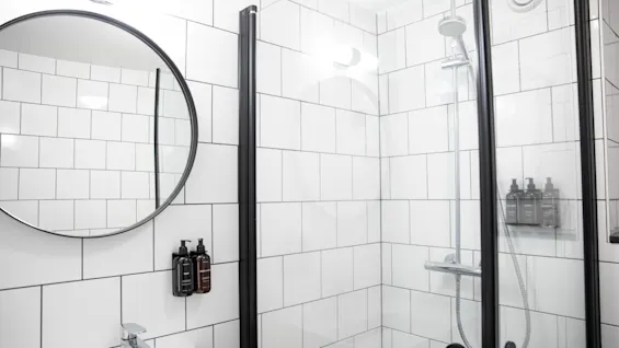 Et rundt spejl hænger på en flisebelagt badeværelsesvæg, der afspejler en moderne brusekabine med glasdøre. Toiletsager er monteret ved siden af vasken, hvilket fremhæver det stilrene, minimalistiske design.