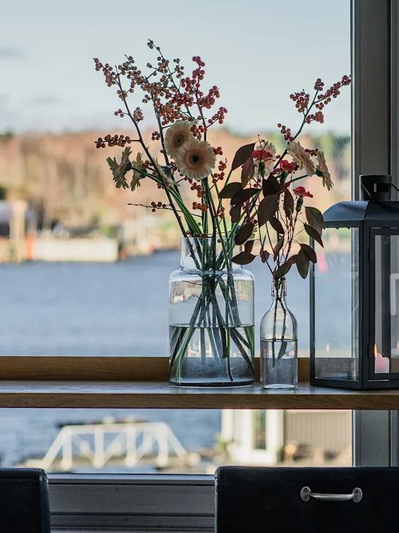 En glasvas med blommor och en svart lykta står på en fönsterbräda med utsikt över lugnt vatten och suddiga träd i bakgrunden.