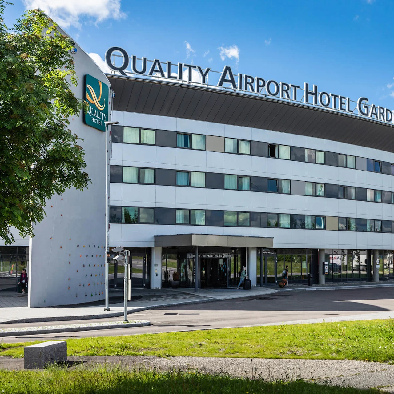 A modern, curved hotel building labeled "Quality Airport Hotel Gardermoen" with a climbing wall, situated amidst trees and flags under a bright blue sky. Text includes "Quality Hotel."