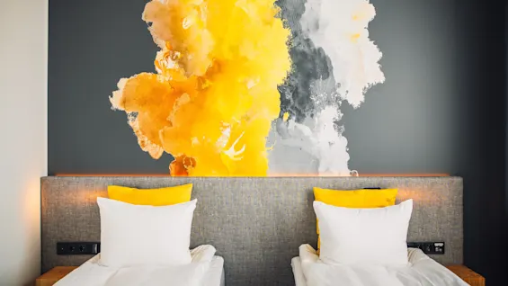 Two beds with white linens and yellow pillows against a dark wall. A large abstract art piece with splashes of yellow, gray, and white decorates the wall, adding vibrant contrast.