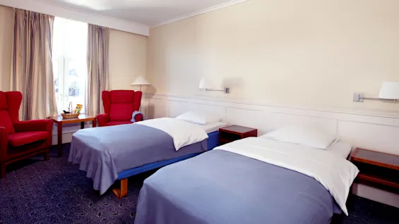Ett hotellrum med två enkelsängar, röda fåtöljer och en öppen balkongdörr.