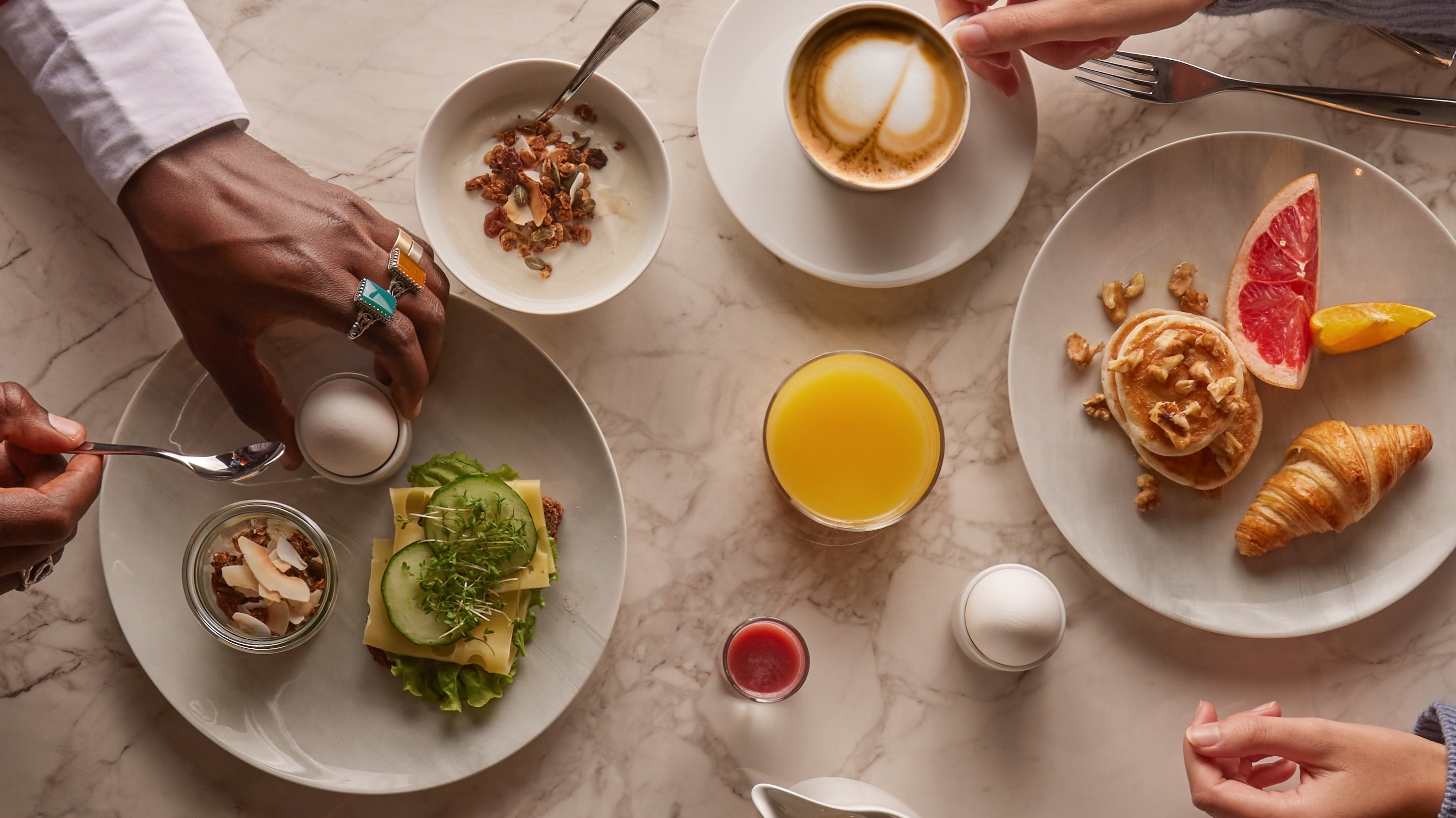 Två personer njuter av en härlig frukostbuffé med kaffe, juice, bakverk och salta rätter på ett marmorbord.