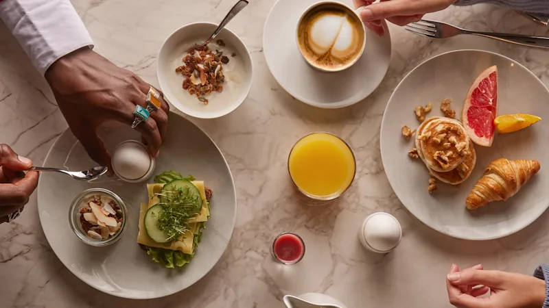 To personer nyder en lækker morgenmadsbuffet med kaffe, juice, bagværk og salte retter på et marmorbord.