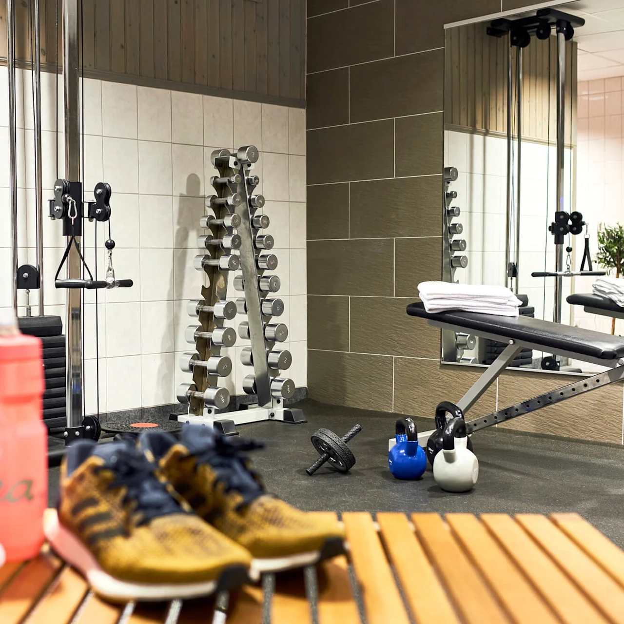 A gym with weights, a bench, towels, a water bottle, and trainers on a wooden slat floor.