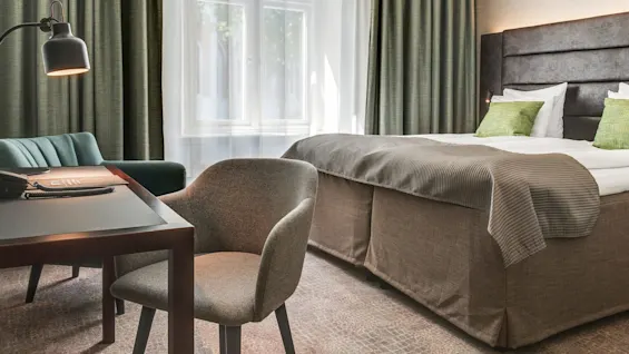 Et velbelyst hotellrom med en seng, arbeidsbord, stoler og store vinduer med grønne gardiner.