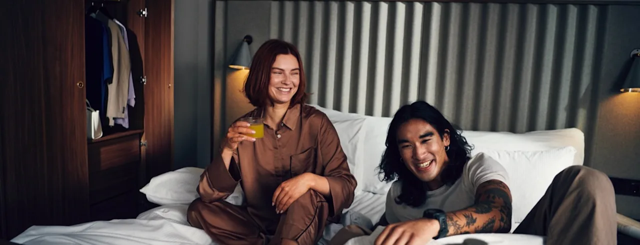 To personer ler og nyter en avslappet frokost på sengen med drikke og mat på et brett.