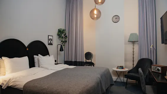 En noggrant bäddad dubbelsäng ligger under två runda pendellampor, omgiven av grå gardiner, en väggklocka och inramad konst i ett mysigt, elegant inrett rum.
