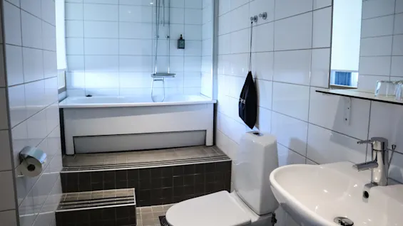 Ett badrum med vita kakelväggar har ett badkar med duschmunstycke. I närheten finns en toalett och ett handfat. En svart väska hänger på väggen; text: ”L’Oréal”.