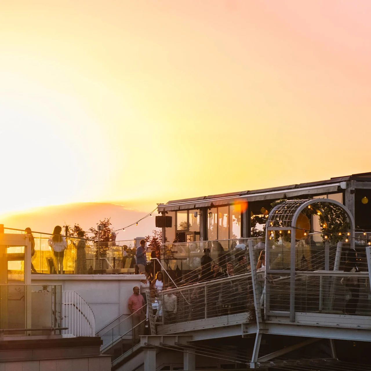 People gather on a rooftop terrace at sunset, with a church spire silhouetted in the background and string lights creating a warm ambiance.