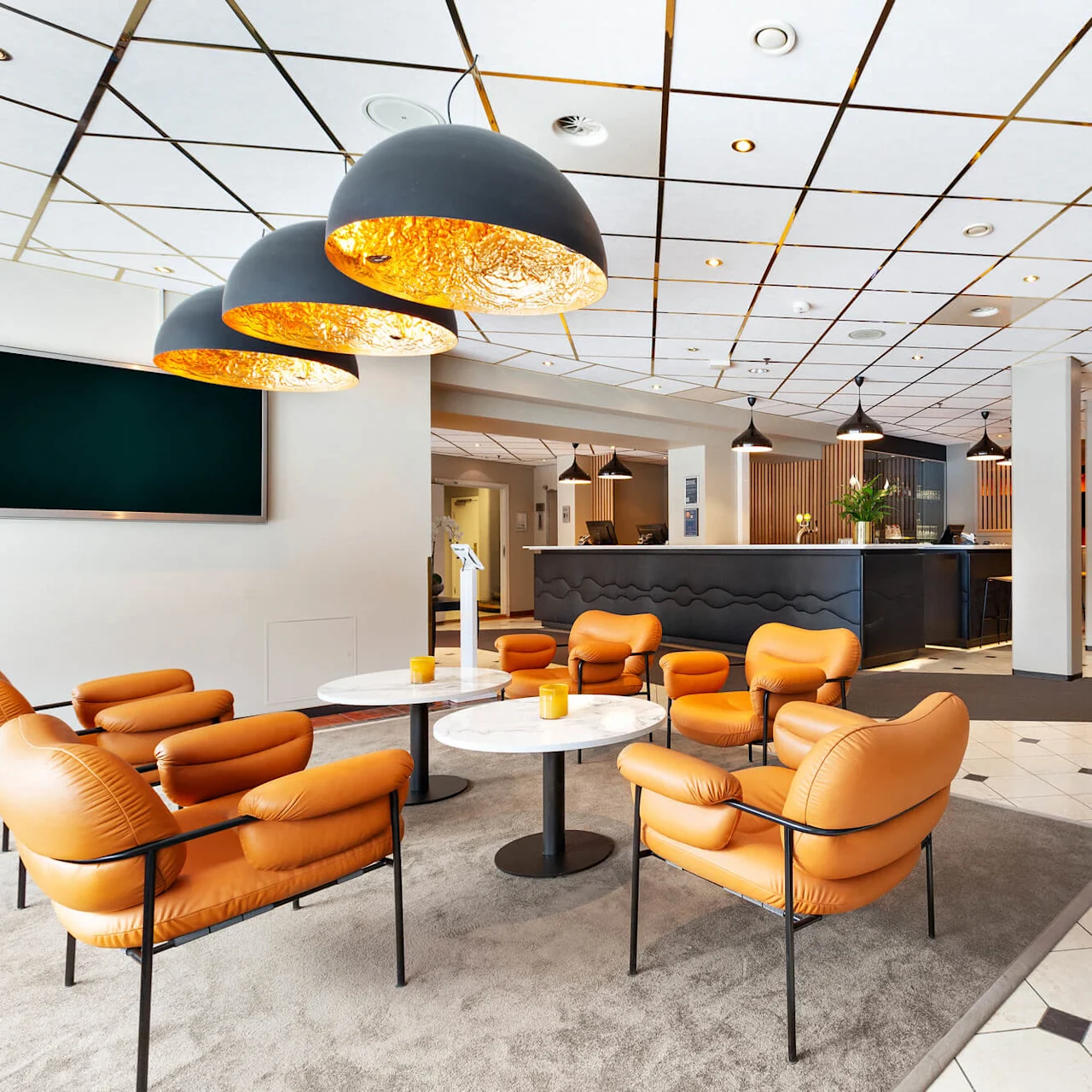 Orange lænestole omkranser runde borde på et tæppe, under store pendellamper. I nærheden står en mørk receptionsskrivebord i en lys, moderne lobby med mønstret loft og flisegulv.