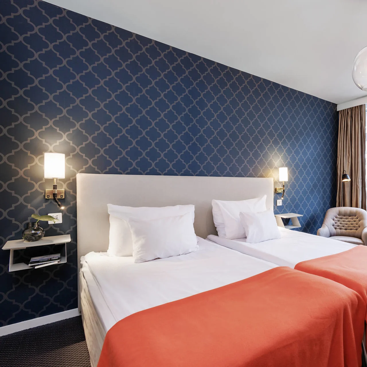 Ett snyggt presenterat hotellrum med mönstrad blå tapet, en stor säng med vitt bäddset och ett orange överkast, sidobord och lampor vid sängen, samt bekväma sittplatser nära ett fönster med gardiner.