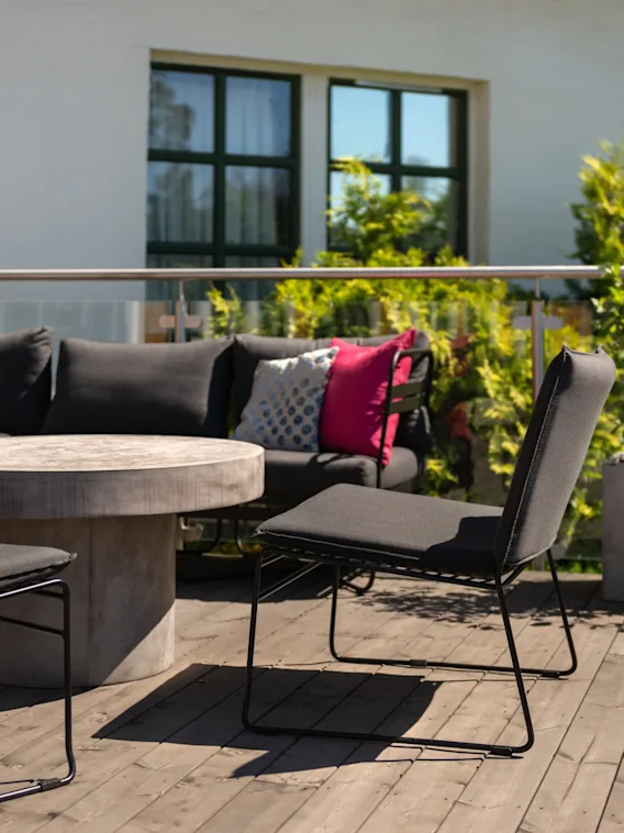 Outdoor seating set surrounds a round stone table on a wooden deck, with vibrant cushions and potted plants against a building backdrop.