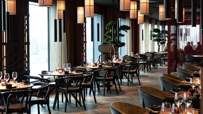 Restaurantborde er elegant dækket med vinglas og bestik, omgivet af moderne indretning og varme hængende lys. Store vinduer giver naturligt lys, mens dekorative træer forbedrer rummet.