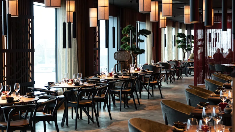 Restaurant tables are elegantly set with wine glasses and cutlery, surrounded by modern decor and warm hanging lights. Large windows provide ambient natural light, with decorative trees enhancing the space.