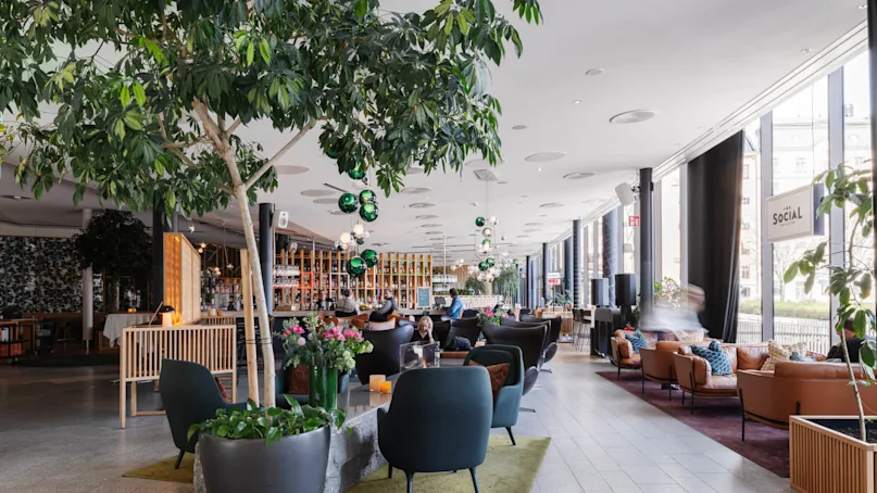 A cozy, modern lounge features people sitting on dark chairs at tables, surrounded by large plants, colorful flowers, and a bar. The sign reads "The Social Market."
