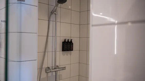 Duschhuvud monterat på en kaklad vägg, med en handhållen dusch fastsatt. Tre mörka flaskor är prydligt placerade bredvid. Miljön är ett modernt badrum med vita kakel.