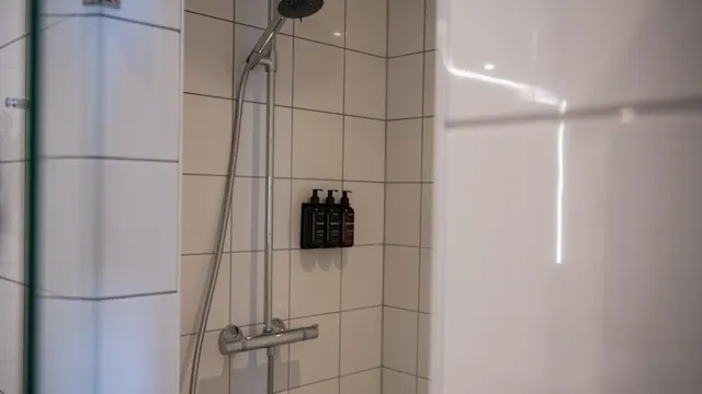 Showerhead mounted on a tiled wall, with a handheld spray attached. Three dark bottles are neatly arranged beside it. The setting is a modern bathroom with white tiles.