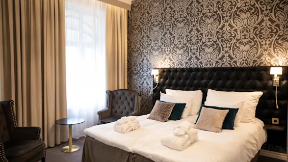 Et hyggeligt hotelværelse med to senge, en mønstret accentvæg og komfortable lænestole ved et vindue.