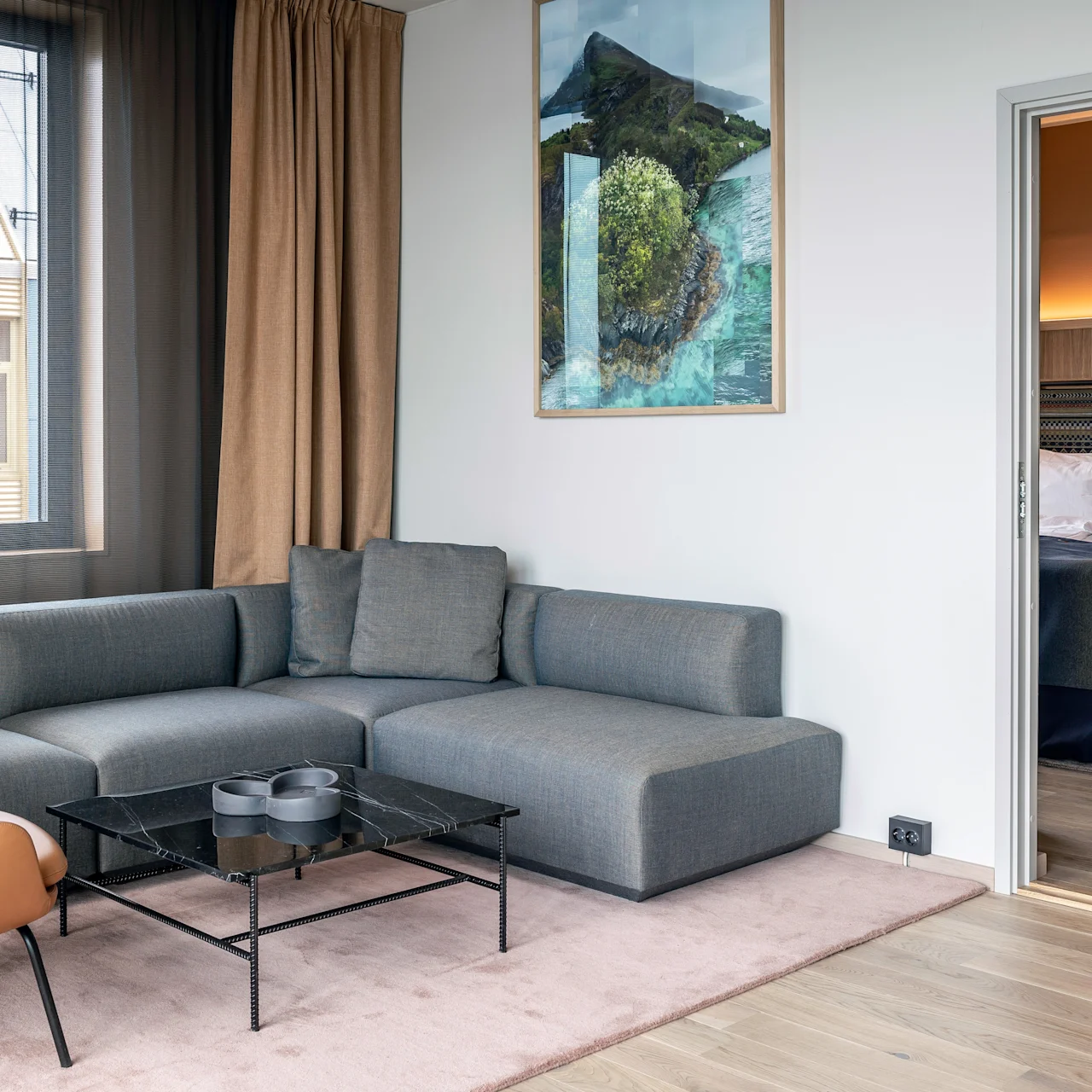 En grå hjørnesofa står i en godt oplyst stue ved siden af en orange stol og et sort sofabord. Et delvist synligt soveværelse med opredt seng ligger ved siden af.