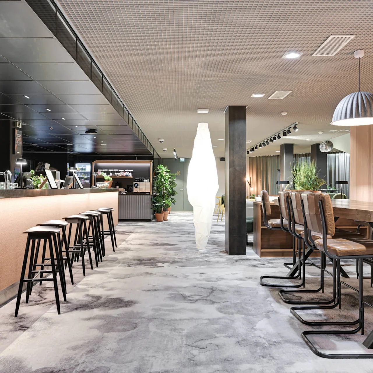 En modern hotellounge har en elegant bar med höga pallar, ett långt gemensamt bord och elegant inredning. Växter ger grönska. Text på väggen: "COMFORT HOTEL XPRESS."