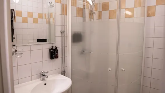 En hvit vask med kran er montert på en flislagt baderomsvegg ved siden av en dusjkabinett i glass. Såpeholdere er festet på veggen; et speil henger over vasken.