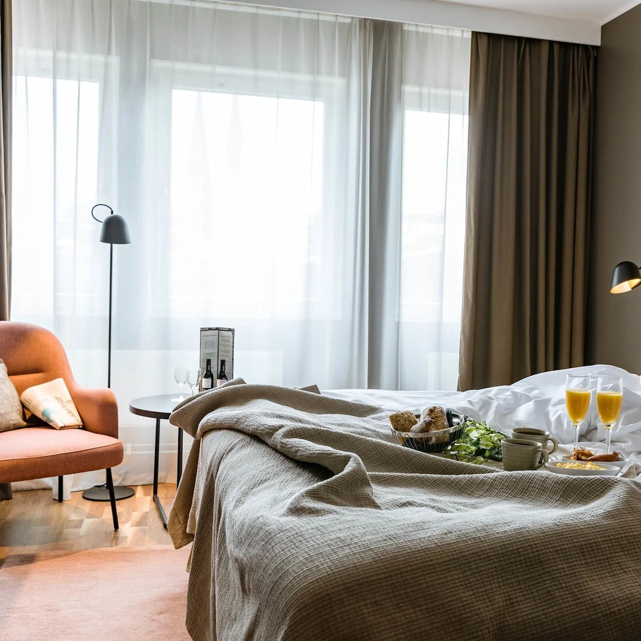 Bed with breakfast tray, containing croissants and orange juice, in a well-lit, cozy hotel room with a beige headboard, brown curtains, and an orange armchair by the window.