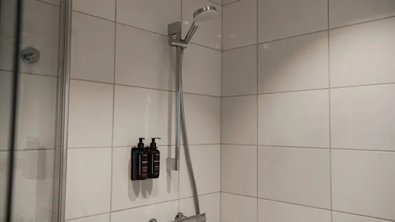 En modern hotell dusch med vita kaklade väggar, ett krom duschhuvud och väggmonterade toalettartiklar, vilket antyder en uppfriskande upplevelse.