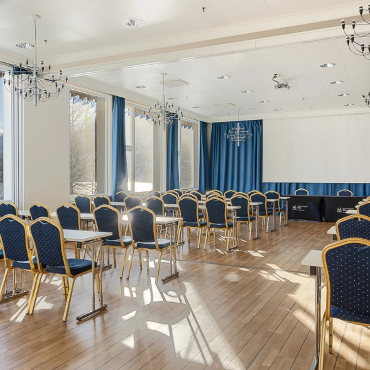 Conference room with rows of blue chairs and tables, facing a large projection screen. Bright sunlight filters through tall windows with blue curtains, illuminating the wooden floor.
